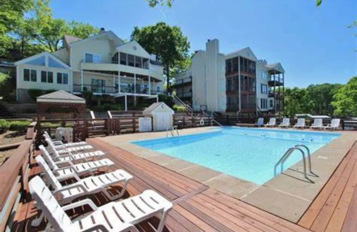 A vibrant outdoor swimming pool surrounded by lounge chairs, with lush trees and resort buildings in the background. The area is spacious, inviting, and clearly designed for families to relax, play, and socialize.