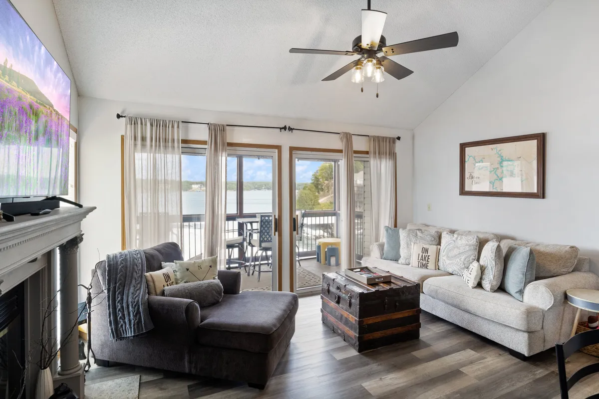 A comfortable living space with a plush sofa, decorative pillows, a TV displaying artwork, and large windows offering sweeping lake views. The room feels inviting and ideal for both social gatherings and quiet relaxation, with the balcony just beyond.