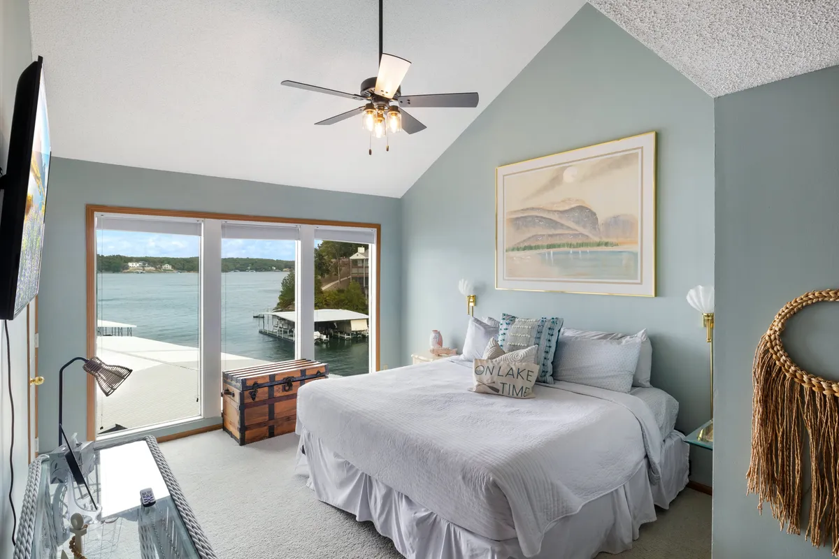 A serene master bedroom featuring a plush bed adorned with decorative pillows, large windows that offer sweeping lake views, and a wooden chest at the foot of the bed. The natural light fills the room, enhancing its tranquil atmosphere and hinting at direct balcony access.