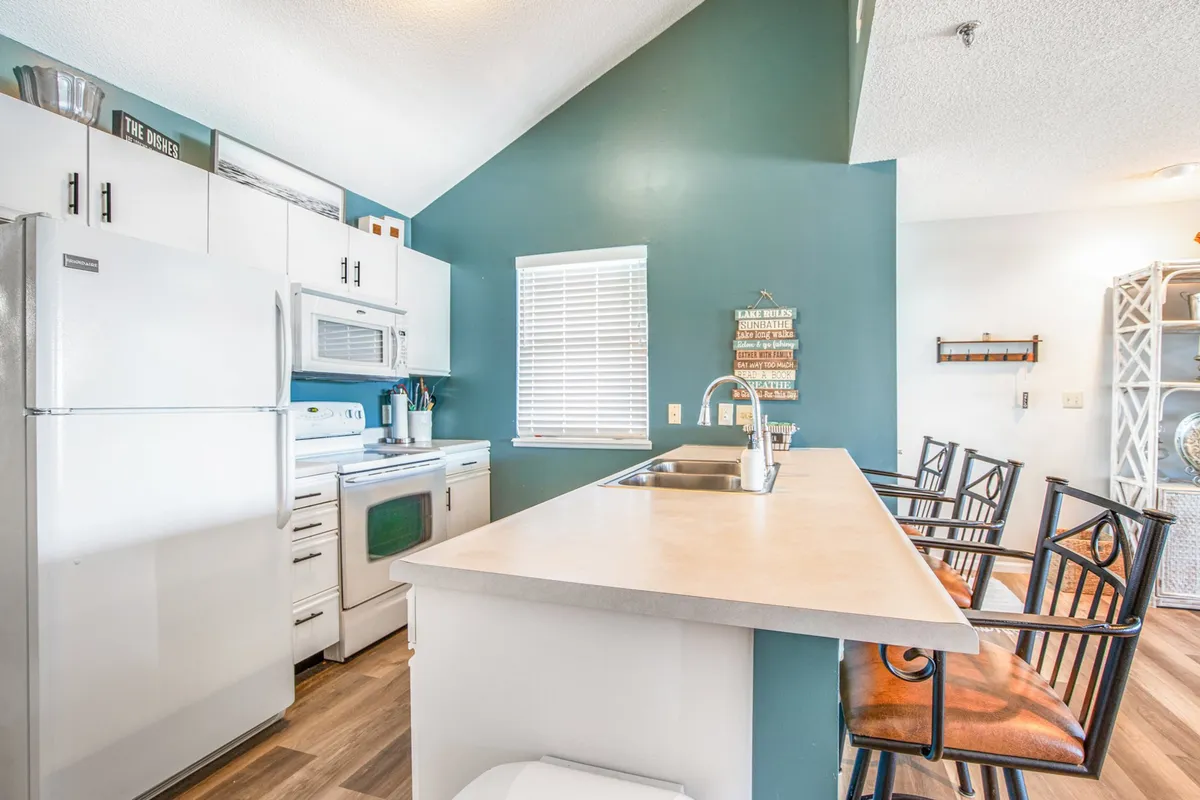 This image features a cheerful and welcoming kitchen with a prominent island, modern stove, refrigerator, a window allowing natural light, and decorative wall art. The layout suggests a space designed for both cooking and socializing, with plenty of workspace and amenities.