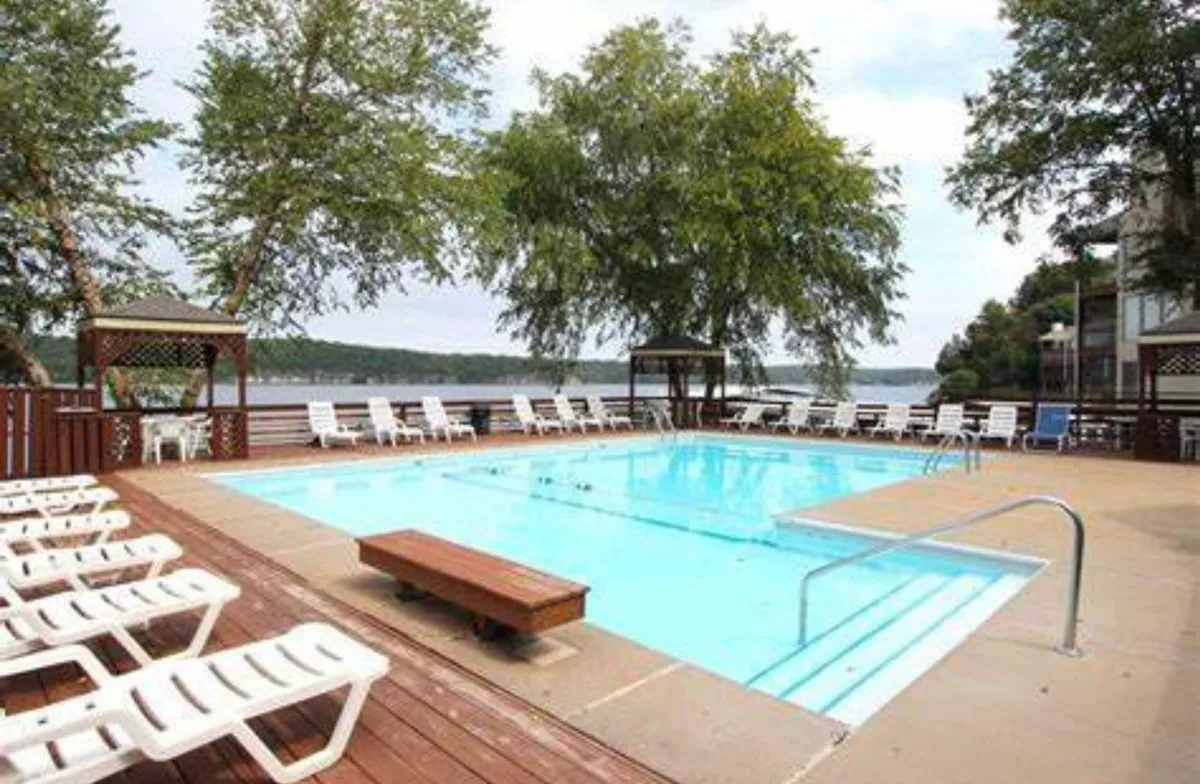 This shot features the tranquil community pool with surrounding trees, lounge chairs for sunbathing, and a cozy gazebo. The lake is visible beyond, reinforcing the pool's proximity to the water, and the atmosphere is peaceful and leisurely.