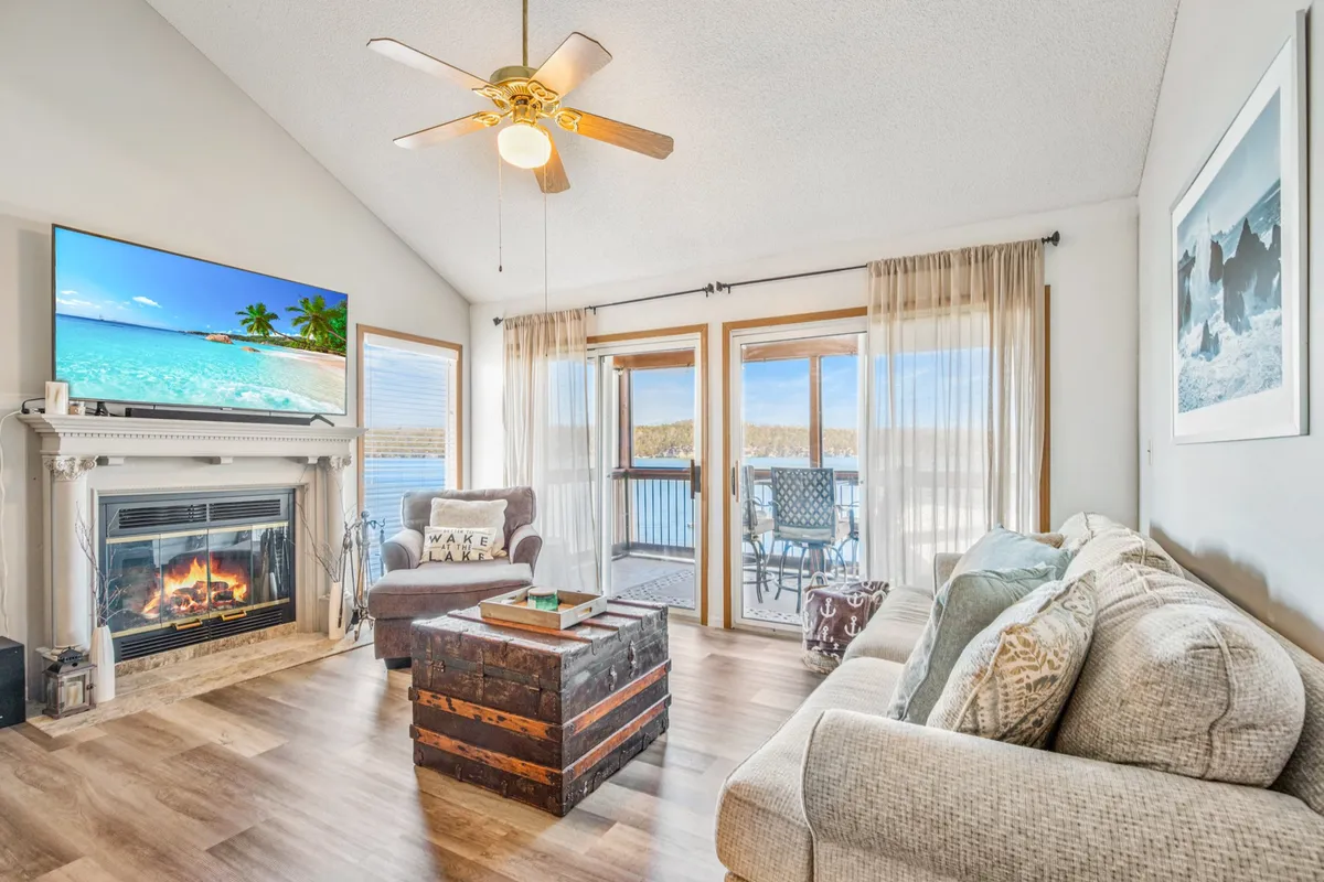This image depicts the central living room, featuring a cozy fireplace, a large smart TV, a roomy coffee table, and large windows that open up to sweeping views of the lake. The space is arranged with comfortable seating, inviting gatherings and relaxation, and the expanse of water visible through the windows draws the outdoors in.