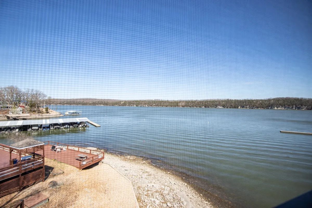 A calm lake surface is seen from the edge of a pier, surrounded by lush trees. The tranquil mood hints at activities like boating, fishing, and enjoying the serenity of nature, with potential for sunset moments and lakeside relaxation.
