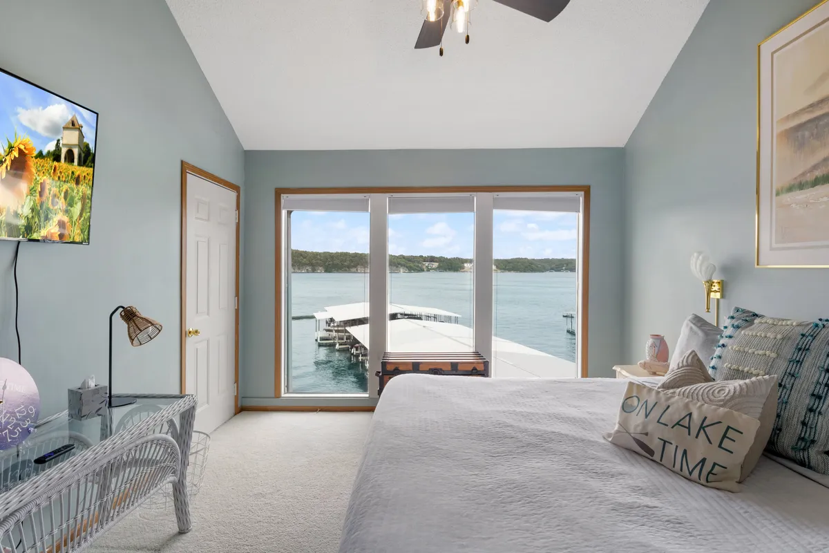 A tranquil queen bedroom with large windows overlooking the lake, a comfortable bed with decorative pillows, and a modern television. The light streaming in underscores the sense of peaceful retreat and the connection to the outdoors.