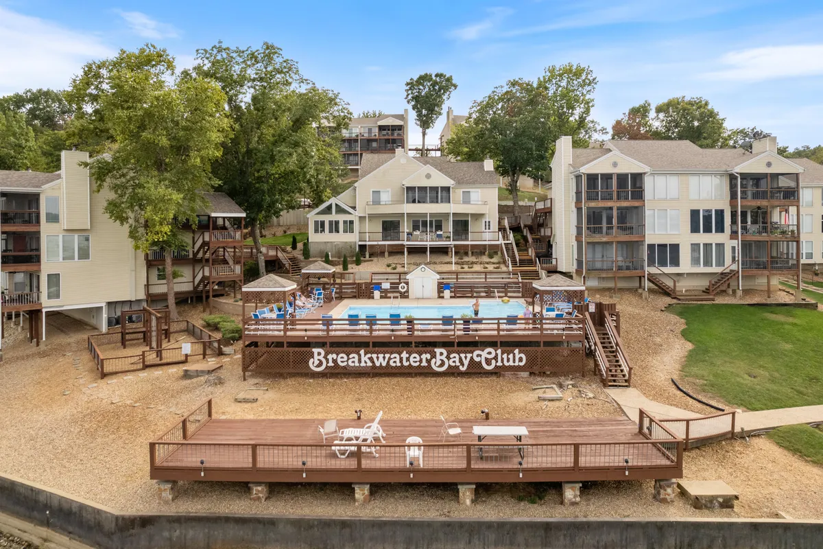 A relaxed exterior view highlighting the swimming pool, the Breakwater Bay Club sign, a deck area for gatherings, and abundant greenery. The setting is perfect for social events or family reunions, with the pool and lake both in sight.