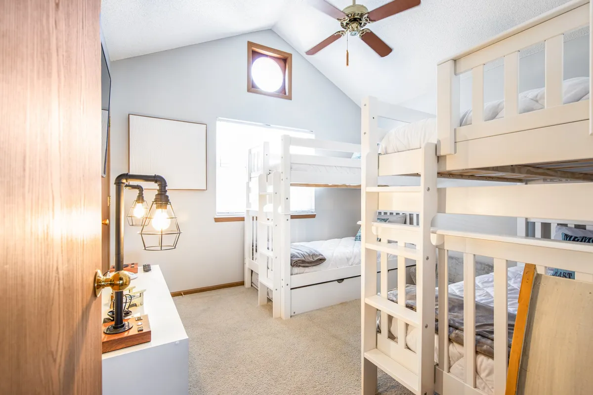 A friendly, relaxed bunk room with sturdy bunk beds, a unique round window, ceiling fan, and a decorative lamp. The room’s playful ambiance is perfect for kids or friends, inviting bonding and fun.