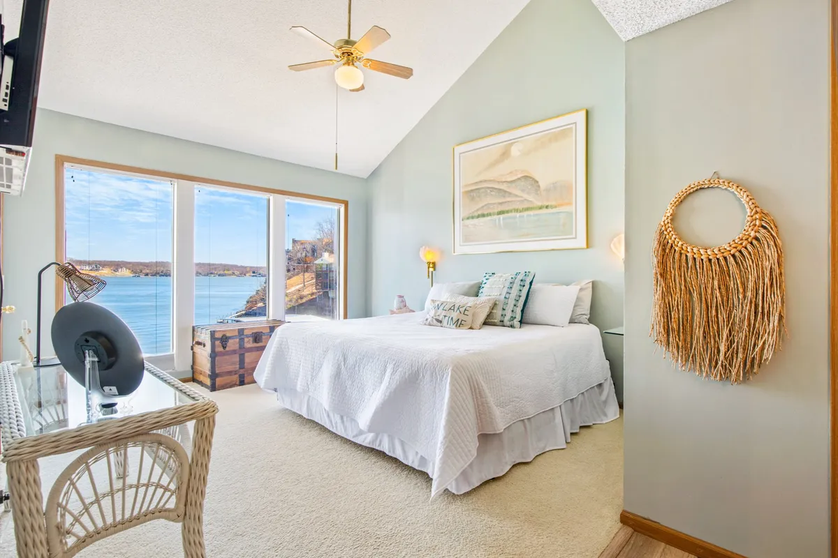The image showcases a sunlit bedroom interior with expansive windows framing a breathtaking lake view. A plush bed with crisp white bedding invites relaxation, while a cozy wicker chair and desk provide the perfect nook for reading, working, or simply soaking in the scenery. Decorative wall art adds a stylish touch, and the space exudes tranquility and comfort. The serene mood is enhanced by natural light spilling across the room, highlighting the seamless connection between indoor comfort and outdoor beauty.